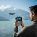Drone DJI Neo 2 Fly More Combo gris con cámara de 12MP y grabación 4K siendo operado mediante control remoto por un hombre frente a un lago con montañas nevadas de fondo
