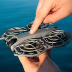 Drone DJI Neo 2 Fly More Combo gris con cámara de 12MP y grabación 4K con botón de encendido siendo activado manualmente mientras se sostiene sobre la palma de la mano fondo azul